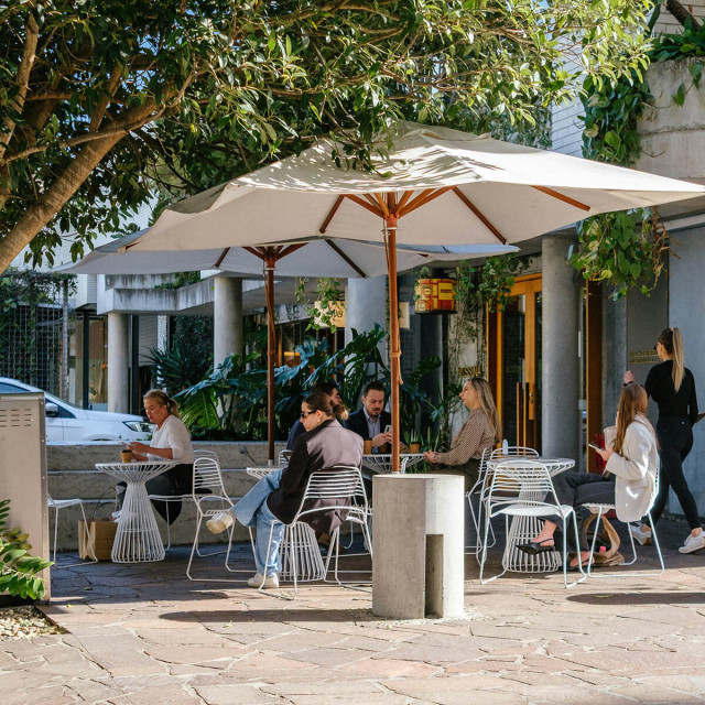 Mehrere Personen sitzen auf der Terasse eines Ladens vor dem Greenhouse, trinken Kaffee und unterhalten sich.