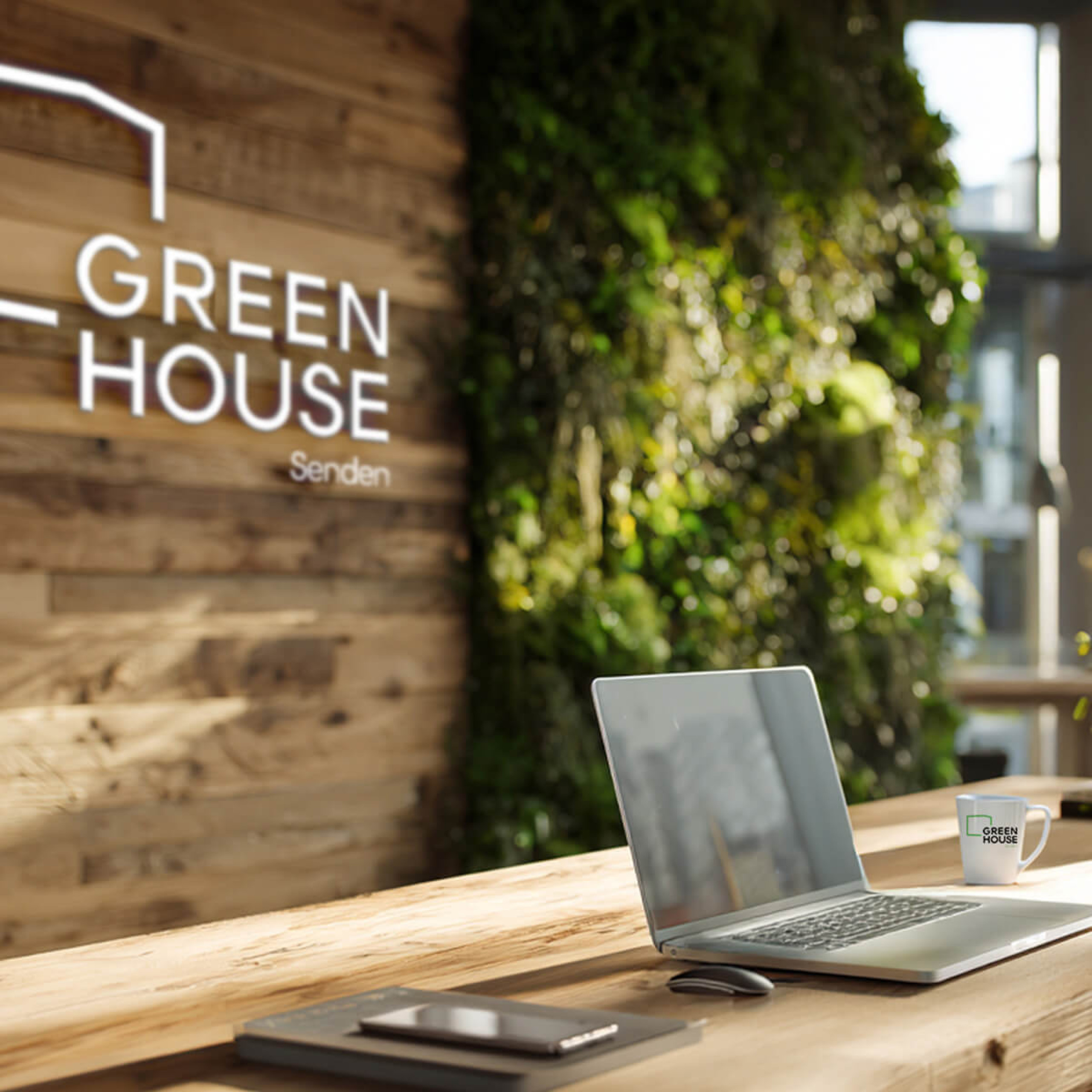 Ein Laptop steht auf einem Tisch und im Hintergrund befindet sich das Greenhouse Senden Logo auf eine zum Teil begrÃ¼nten Wand.